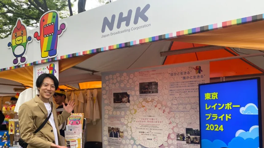 An outdoor NHK festival booth adorned with rainbow borders and a rainbow Domo-kun mascot. A man smiles and waves in front of a large message board filled with written notes, next to a blue digital screen reading "Tokyo Rainbow Pride 2024."