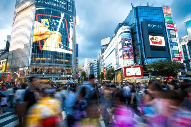 A busy crowd with motion blur crossing the street at the famous Shibuya Crossing intersection in Tokyo, surrounded by bright neon signs and electronic video billboards.