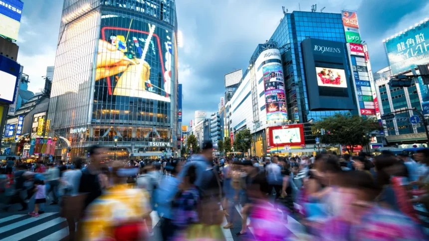 A busy crowd with motion blur crossing the street at the famous Shibuya Crossing intersection in Tokyo, surrounded by bright neon signs and electronic video billboards.
