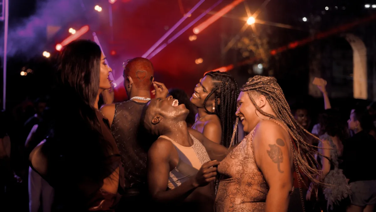 A group of diverse friends laughing and dancing together at an outdoor nighttime party or club event. In the center, a person covered in body glitter laughs with their head tilted back, surrounded by smiling friends with long braids and stylish outfits, set against a dark background illuminated by vibrant red and purple stage lights.