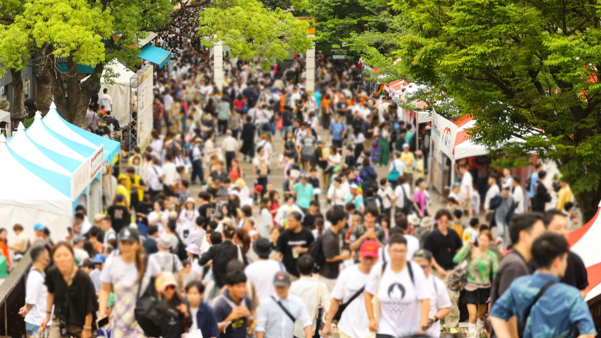 A crowded pedestrian path at the Tokyo Rainbow Pride festival hub in Yoyogi Park. This vibrant outdoor snapshot captures the massive scale of the Tokyo Pride 2025 event, with attendees exploring sponsor booths and tents framed by lush summer greenery.