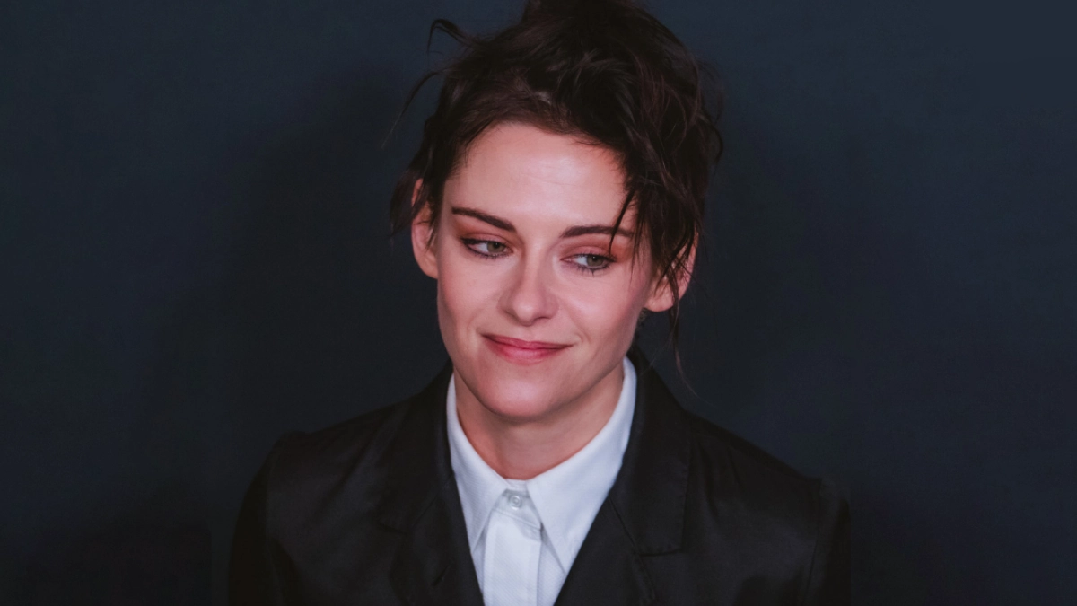 A close-up portrait of Kristen Stewart at the WWD Style Awards, showcasing a refined soft masc aesthetic. She wears a structured black blazer over a buttoned-up white collared shirt, sporting a subtle smile and looking slightly to the side. Her dark hair is styled in a loose, textured, and slightly messy updo against a dark gray backdrop.