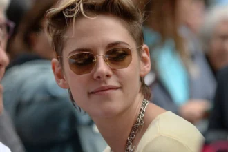 A close-up portrait of Kristen Stewart at a red carpet event, wearing a chain necklace and dark sunglasses.