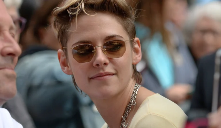A close-up portrait of Kristen Stewart at the WWD Style Awards, showcasing a refined soft masc aesthetic. She wears a structured black blazer over a buttoned-up white collared shirt, sporting a subtle smile and looking slightly to the side. Her dark hair is styled in a loose, textured, and slightly messy updo against a crowd backdrop.