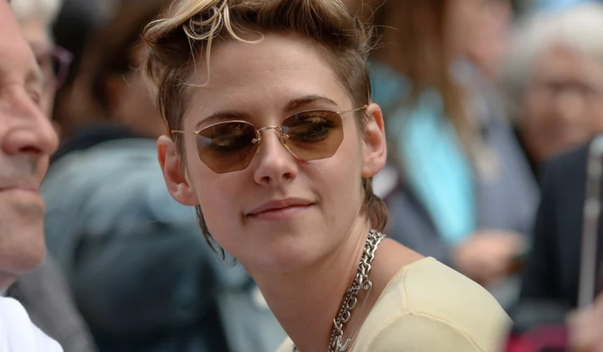 A close-up portrait of Kristen Stewart at a red carpet event, wearing a chain necklace and dark sunglasses.