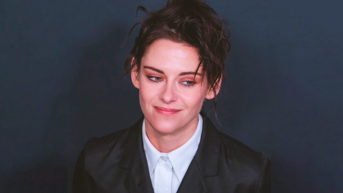 A close-up portrait of Kristen Stewart at the WWD Style Awards, showcasing a refined soft masc aesthetic. She wears a structured black blazer over a buttoned-up white collared shirt, sporting a subtle smile and looking slightly to the side. Her dark hair is styled in a loose, textured, and slightly messy updo against a dark gray backdrop.