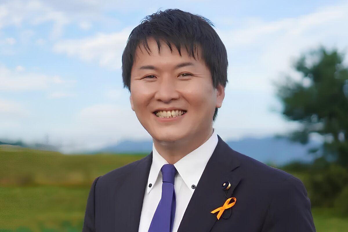 Mayor Toshinao Inamori wearing a purple tie and yellow ribbon lapel, with a grassy field in the background