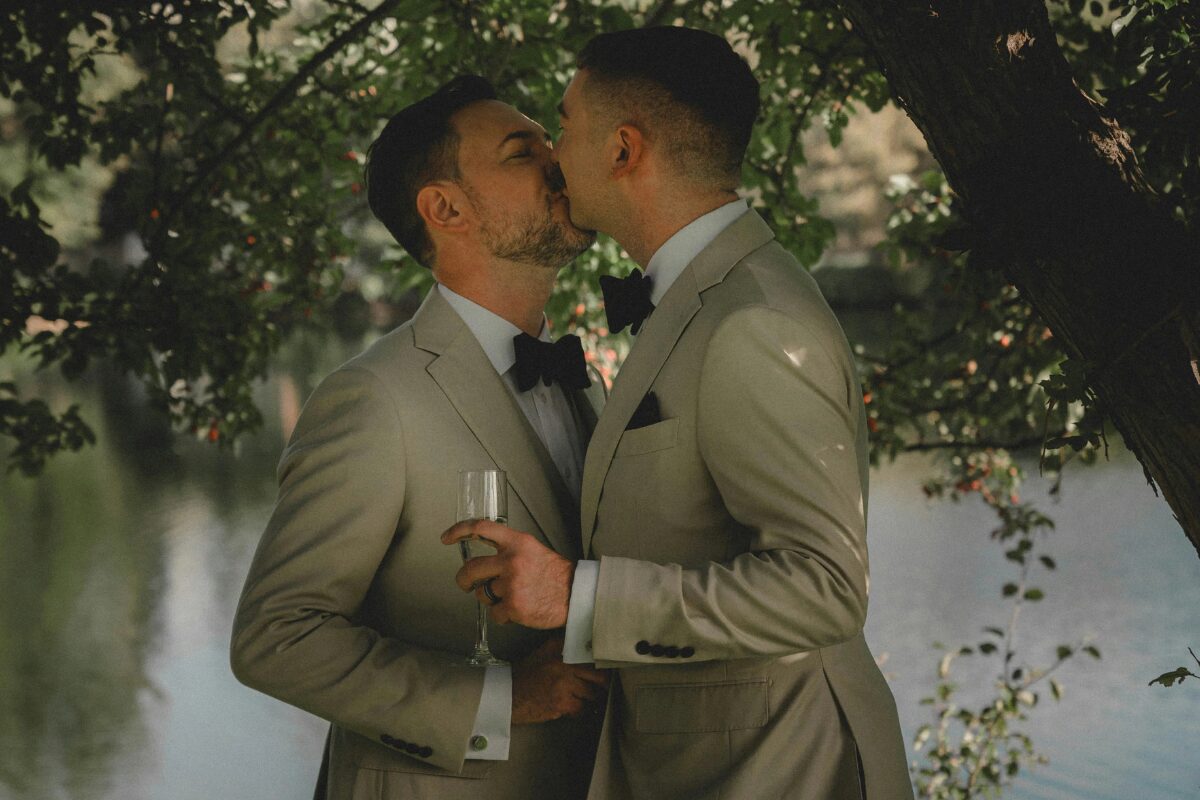 Two men wearing light colored tuxedos and black bowties under a tree have their first kiss as a married couple