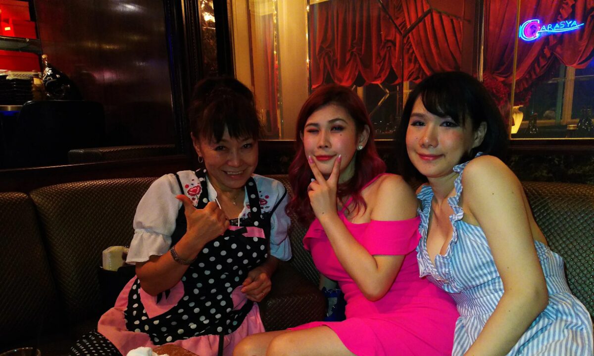 Three women sit at a booth in Garasya, one wearing a black and white polka-dot pinafore on the left, the woman in the middle wearing a pink dress, and the woman on the right wearing a blue and white vertical striped dress
