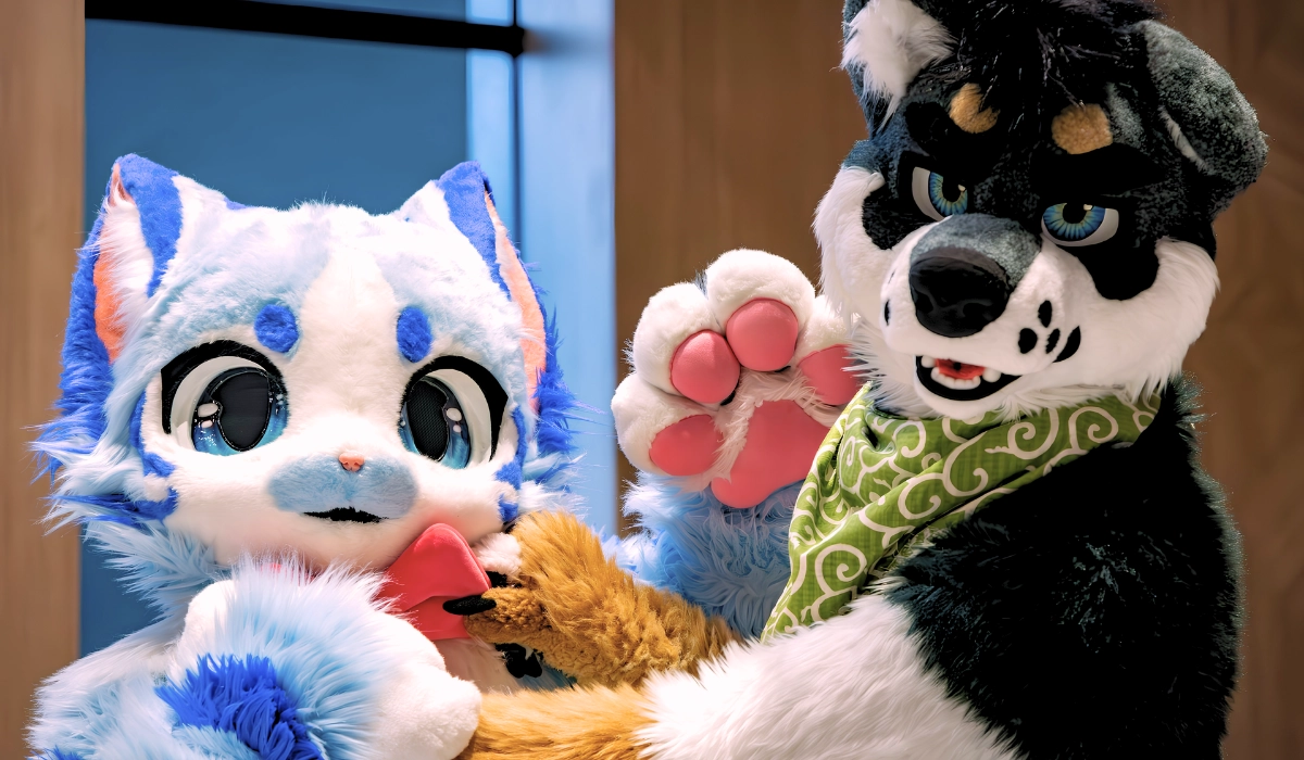 On the left, a kemono fursuit of a light blue and white canine with large anime-style blue eyes. On the right, a black, white, and tan husky fursuit in a western style, wearing a green patterned bandana and waving a white paw with bright pink paw pads.
