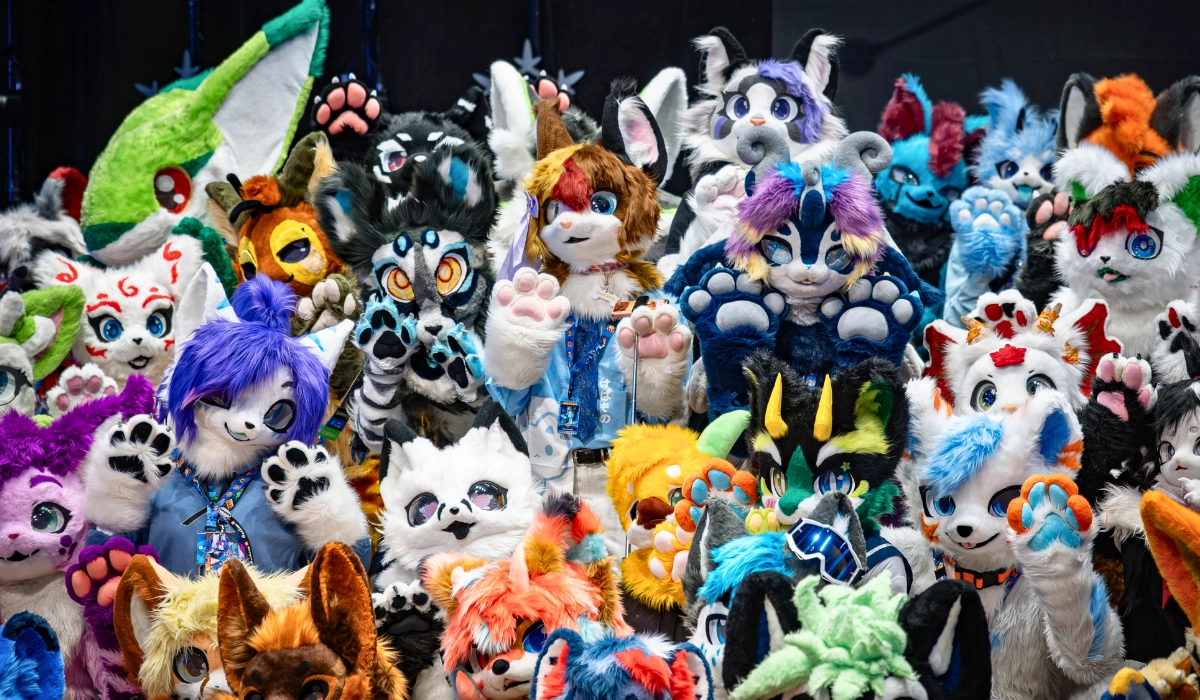 A large group of colorful fursuiters posing tightly together for a group photo, displaying a wide variety of species, bright colors, and kemono fursuit styles with many waving their paws