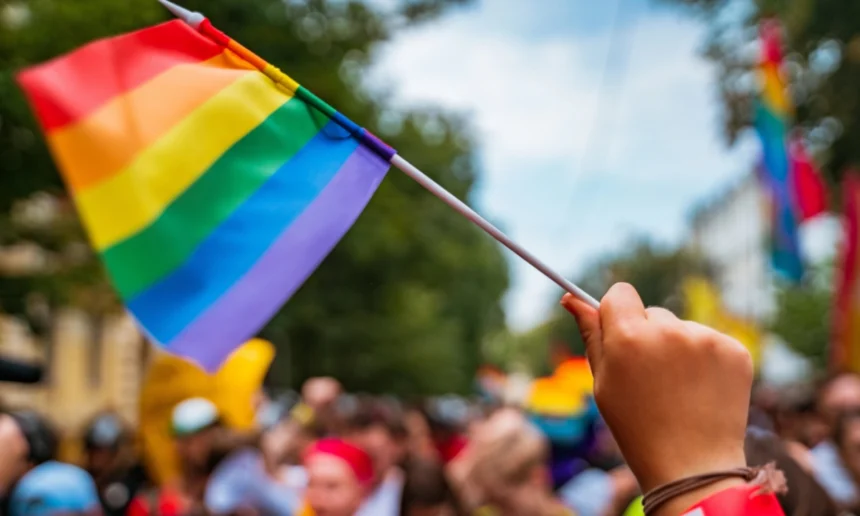 Pride flag is being held into the air and waved at a pride march, while person is surrounded by crowd.