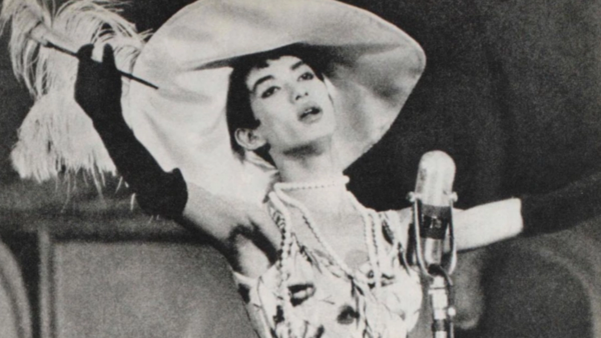 A young Akihiro Miwa poses in front of a microphone mid-song, dressed in extravagant clothing and a pearl necklace. 