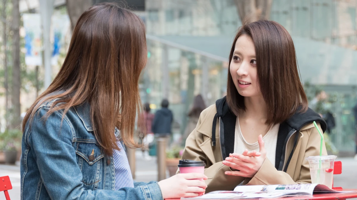 Two Japanese women talk while sat outside drinking coffee.