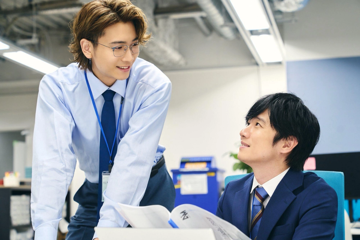 Two Japanese men in office attire smile at each other while talking across a desk in a modern workplace.