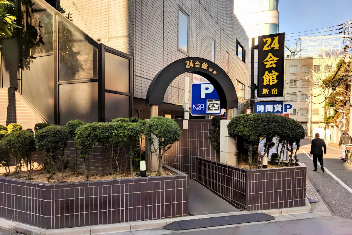 Exterior of 24 Kaikan Shinjuku, showing the building entrance, black-and-gold Japanese signage, greenery, and pedestrians on a Tokyo street.