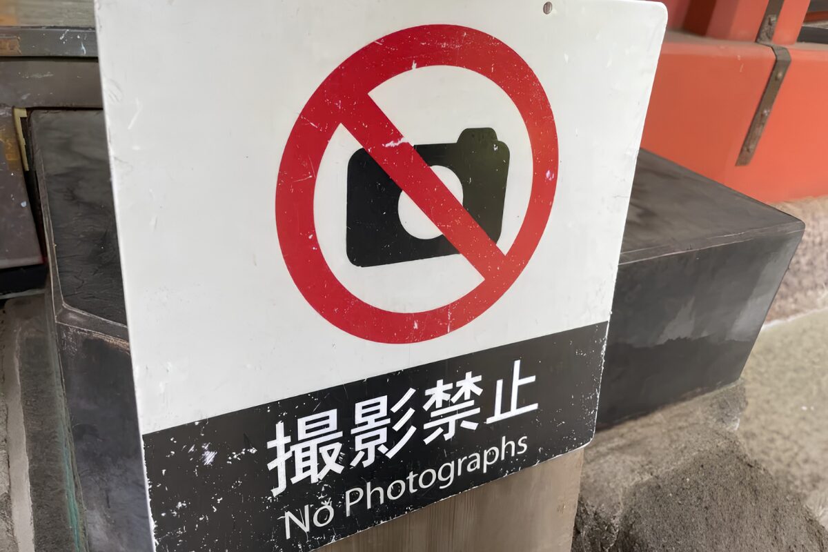 Close-up of a worn “No Photographs” sign in Japan, showing a camera icon crossed out in red with Japanese text reading 撮影禁止.