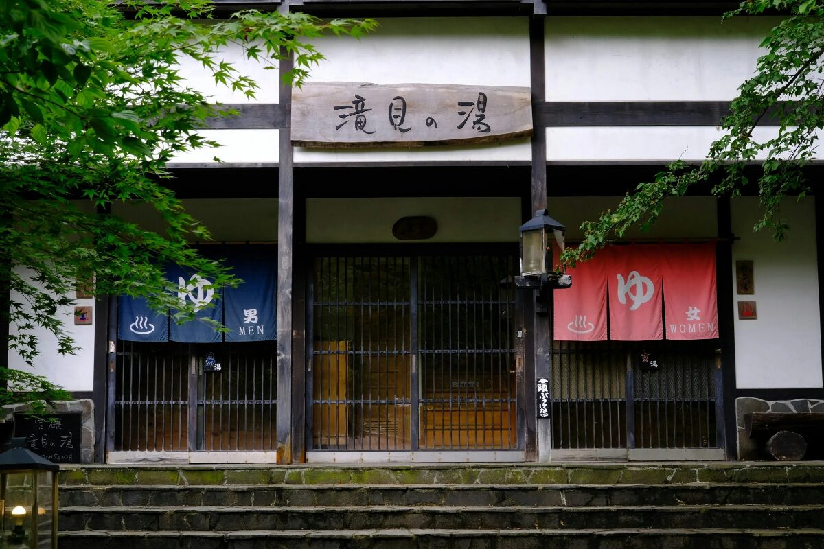 Onsen exterior. There are three doorways: one central one for staff, and one for men on the left and women on the right. The gendered entrances are marked by a blue cloth divider for men, and a red cloth divider for women.