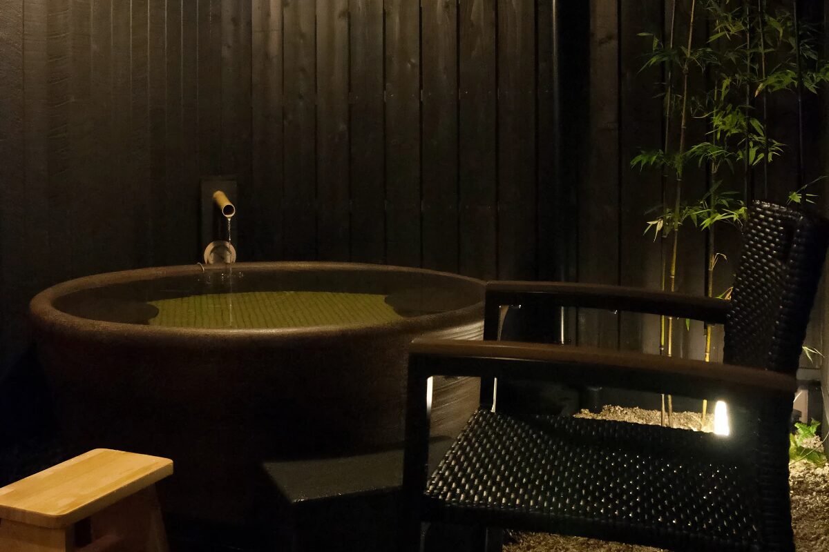A private bath, located outside. Water trickles into a large bowl-shaped bath, with a wicker chair in the foreground