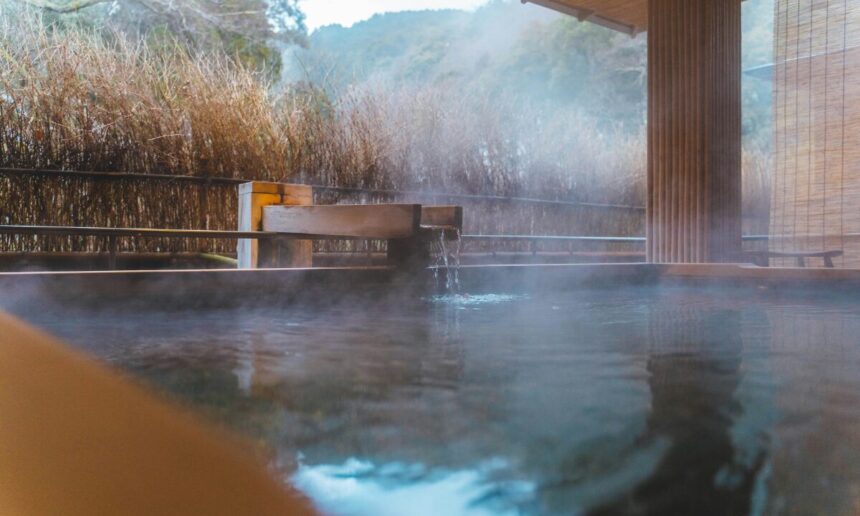 An outdoor onsen bath. In the middle distance, water pours from a wooden spout. Steam rises from the water. In the background is a wooded mountain.