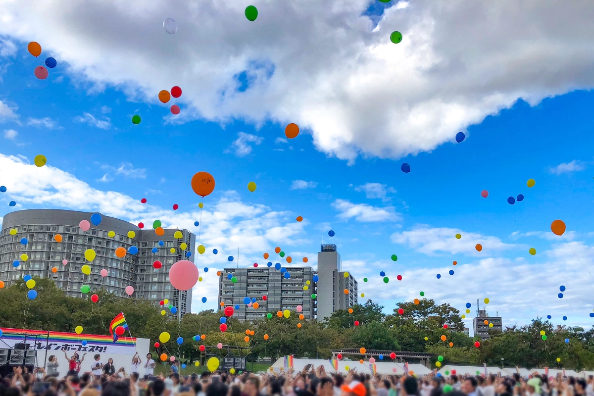Colorful balloons float above a large Pride festival crowd gathered outdoors beneath a bright blue, partly cloudy sky, with a rainbow flag visible near the stage.