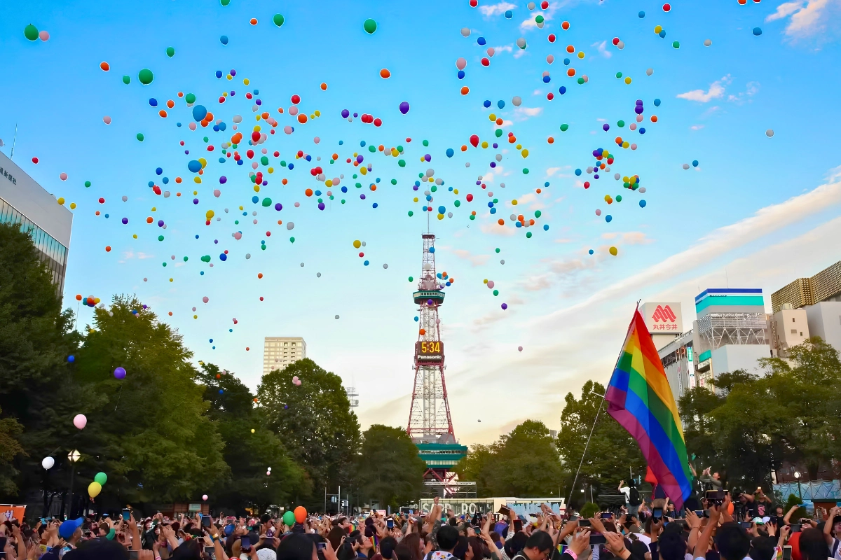 Colorful balloons rise above a large Pride crowd in central Sapporo, with a rainbow flag and the Sapporo TV Tower visible against the evening sky.