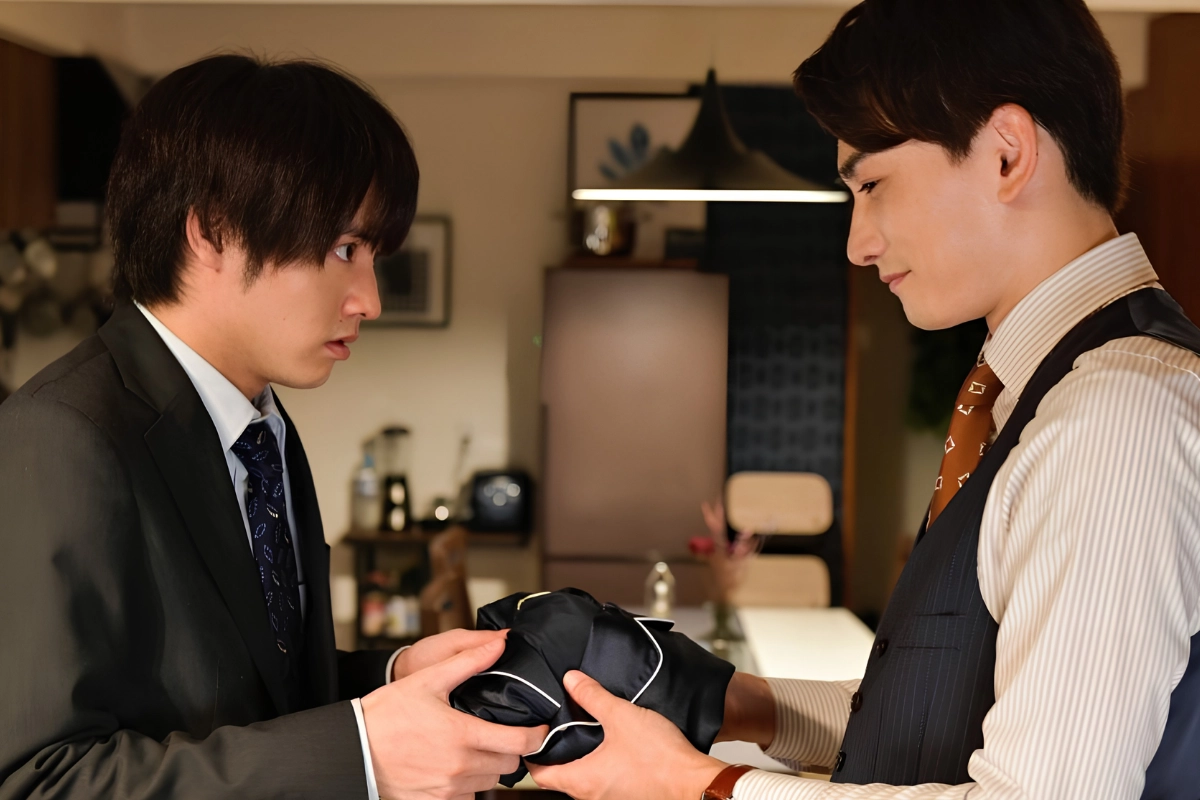 Two Japanese men in formal work clothes exchange a folded black garment while facing each other inside a warmly lit apartment.