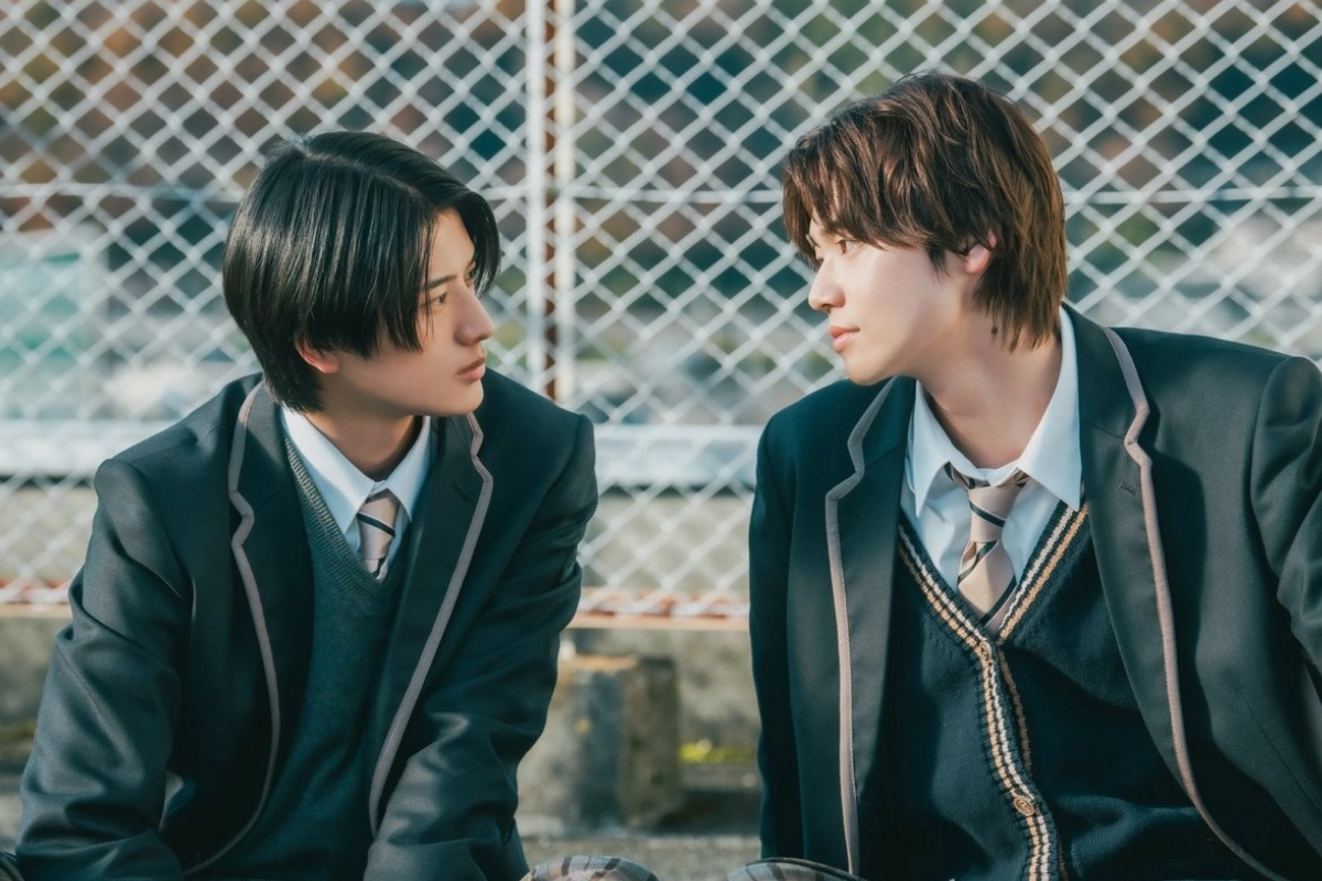 Two Japanese male students in matching school uniforms sit beside each other outdoors, looking intently into each other’s eyes.