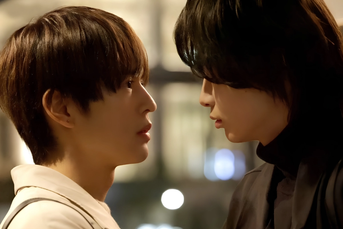 Two young Japanese men stand face-to-face at night, gazing closely at each other in a warmly lit indoor setting.