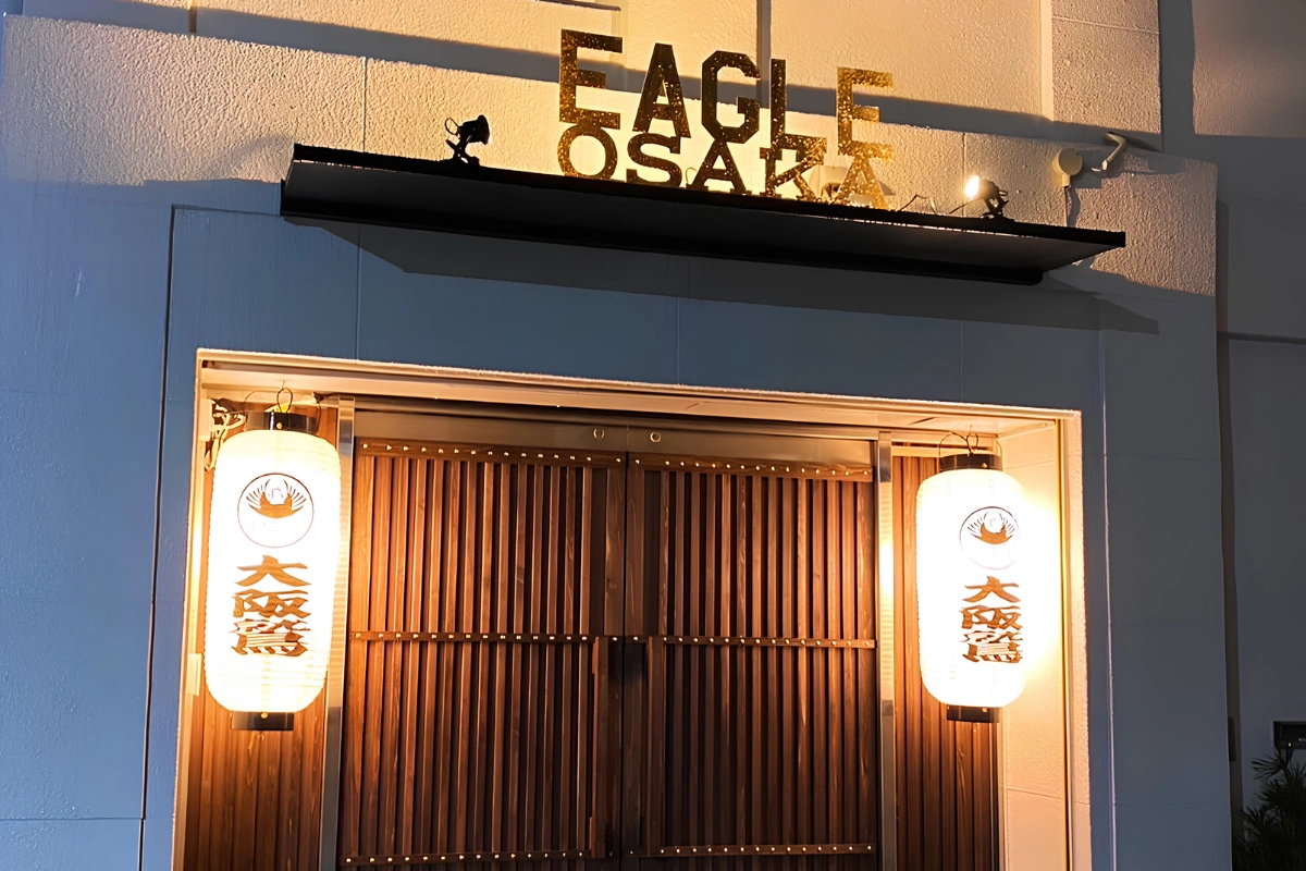 Illuminated entrance to Eagle Osaka, with wooden doors, two glowing Japanese lanterns, and the venue’s name displayed above.