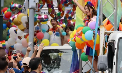 Pride parade participant stands on a decorated float surrounded by colorful balloons and rainbow flags, raising one arm as spectators cheer from the street.