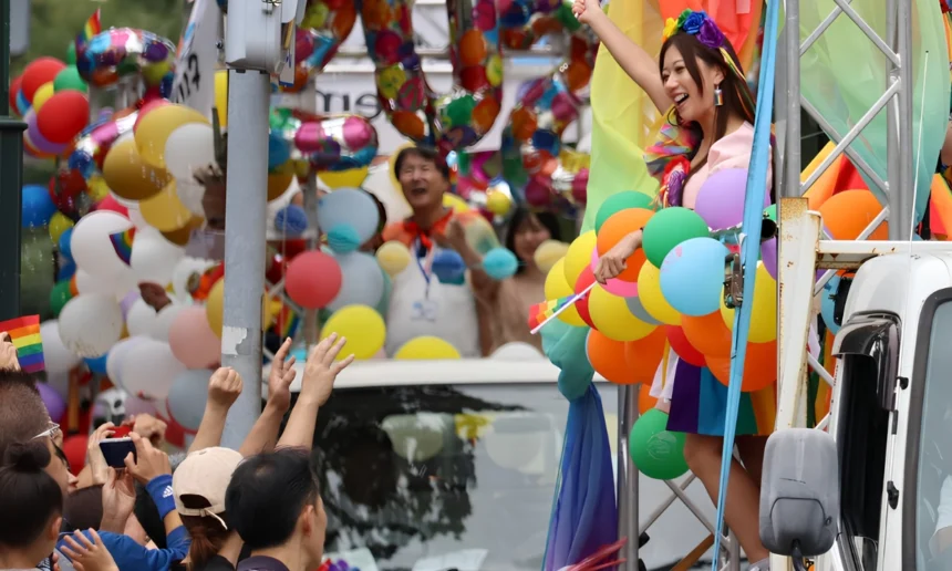 Pride parade participant stands on a decorated float surrounded by colorful balloons and rainbow flags, raising one arm as spectators cheer from the street.