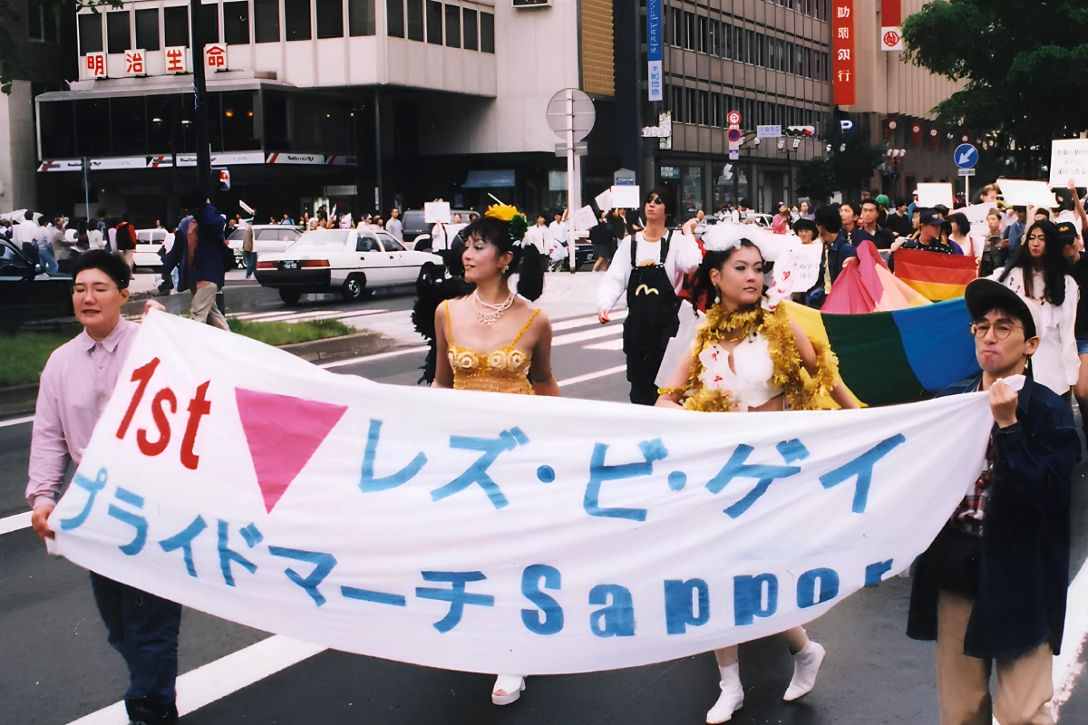 Participants in the first 1996 Sapporo Pride march carry a banner reading “1st Les-Bi-Gay Pride March Sapporo” through a city street.