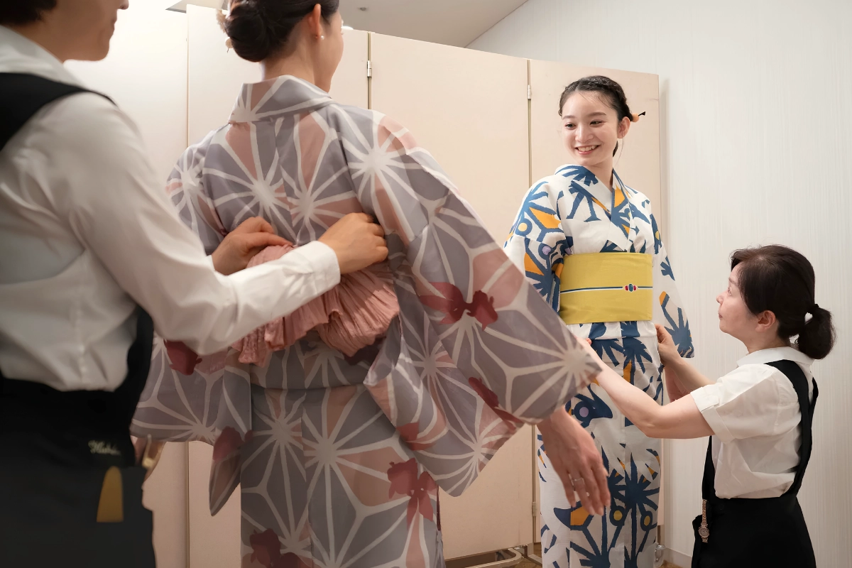 Two kimono dressers adjust the obi and fit of customers wearing patterned yukata in a bright dressing room.