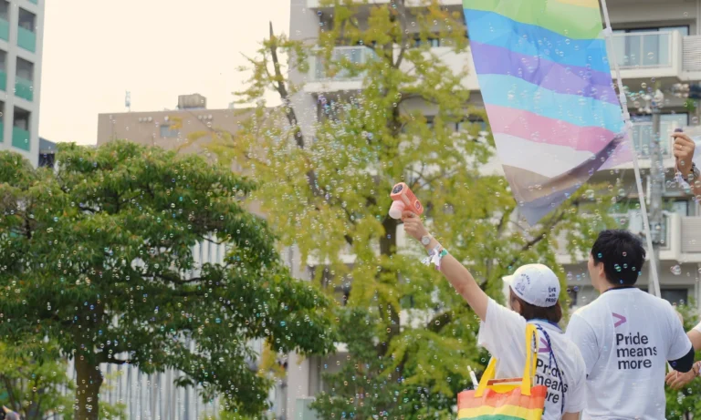 Two people at a Pride event stand beneath a large transgender Pride flag while one raises a bubble gun, filling the air with soap bubbles.