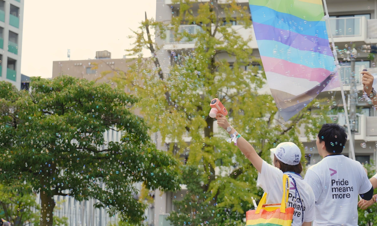 Two people at a Pride event stand beneath a large transgender Pride flag while one raises a bubble gun, filling the air with soap bubbles.