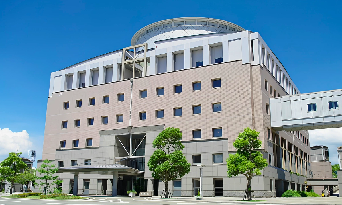 Exterior of a large modern Japanese hospital building under a clear blue sky, with trees lining the entrance and an enclosed walkway connecting to an adjacent wing.