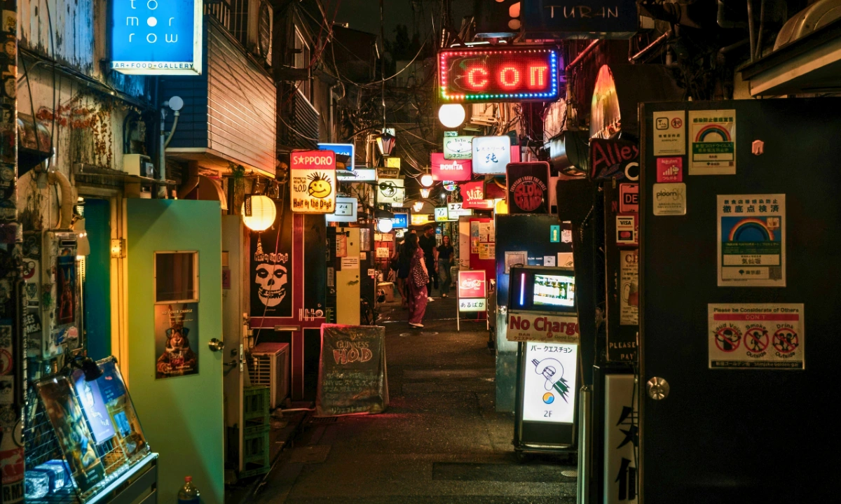 Nighttime view of a narrow Shinjuku Golden Gai alley lined with small bar entrances, illuminated signs, and posted notices.