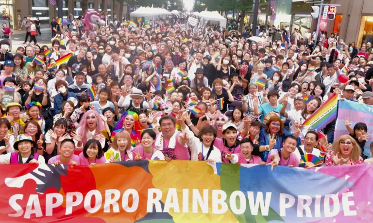 Large Sapporo Rainbow Pride crowd posing behind a colorful event banner, with many participants holding rainbow flags in a busy city-center street.