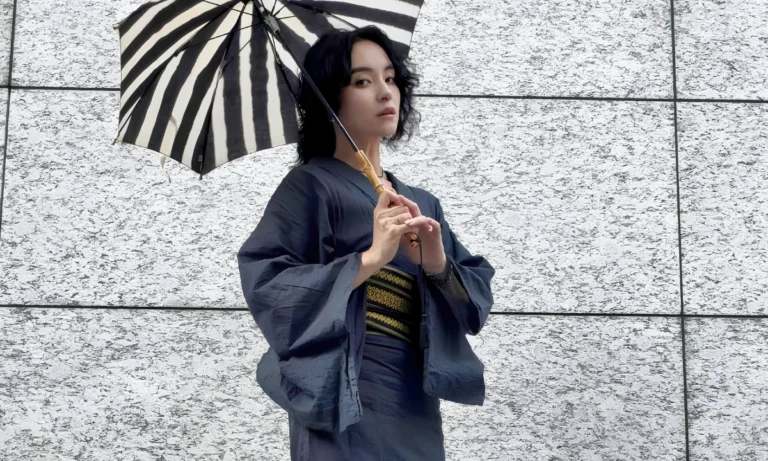 Person wearing a dark contemporary kimono holds a black-and-white striped parasol while standing against a light stone wall.