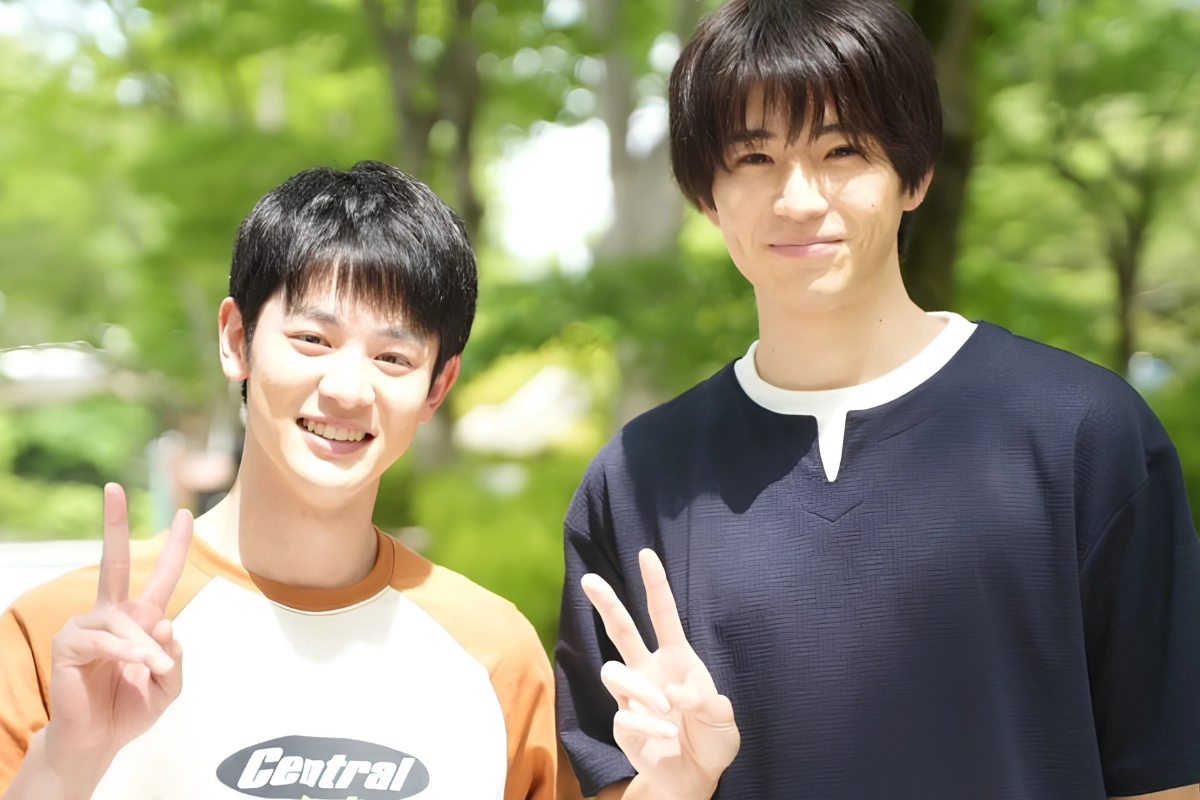 Two young Japanese men smile outdoors in a leafy park, each holding up a peace sign toward the camera.
