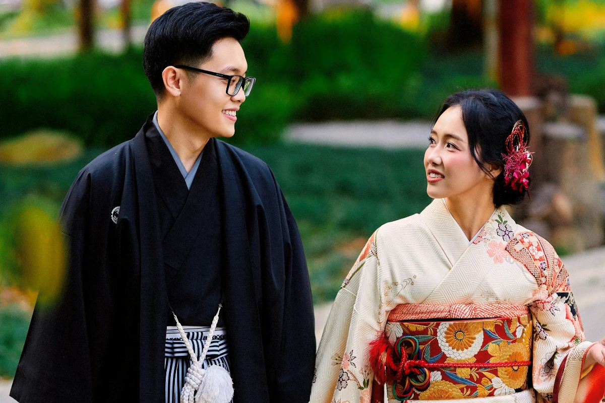 Two people in traditional kimono walk outdoors, with one wearing a dark men’s-style ensemble and the other a light floral kimono with a wide decorative obi.