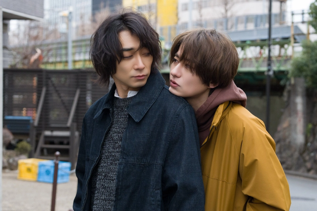 Two Japanese men stand close together on a city street, with one leaning toward the other’s shoulder as they exchange a sideways glance.