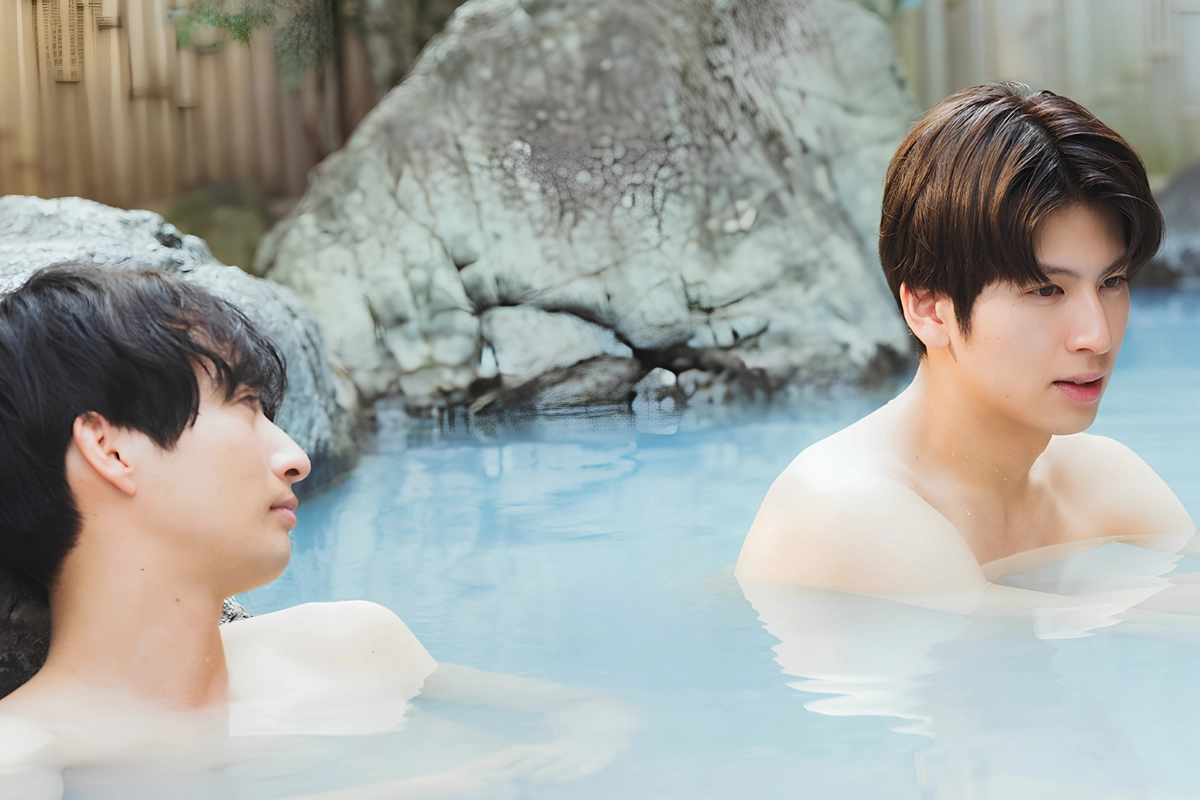 Two Japanese men relax together in an outdoor hot spring, sitting apart in the milky blue water among large rocks.