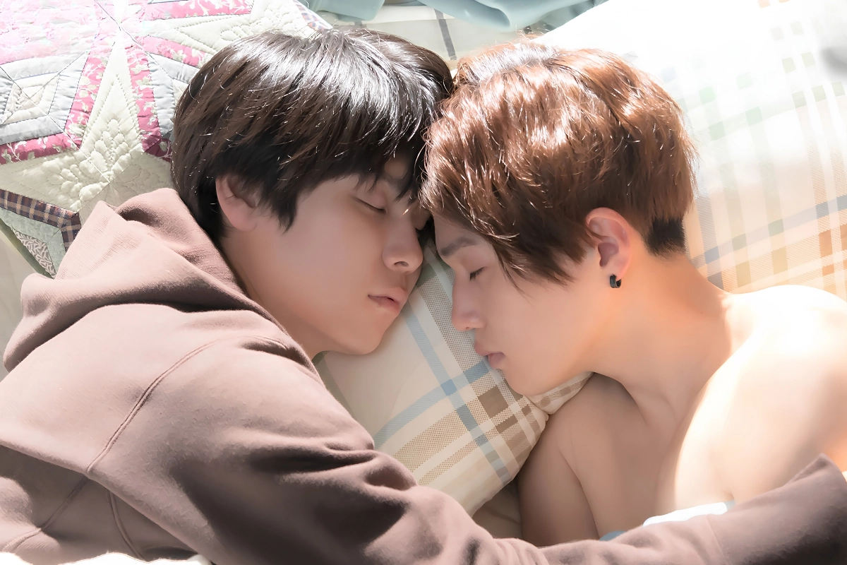 Two young Japanese men lie close together on a bed with their foreheads touching and eyes closed.