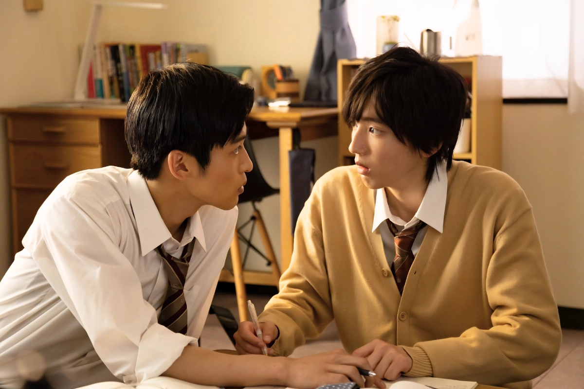 Two Japanese male students in school uniforms sit closely together at a desk, looking at each other while studying.