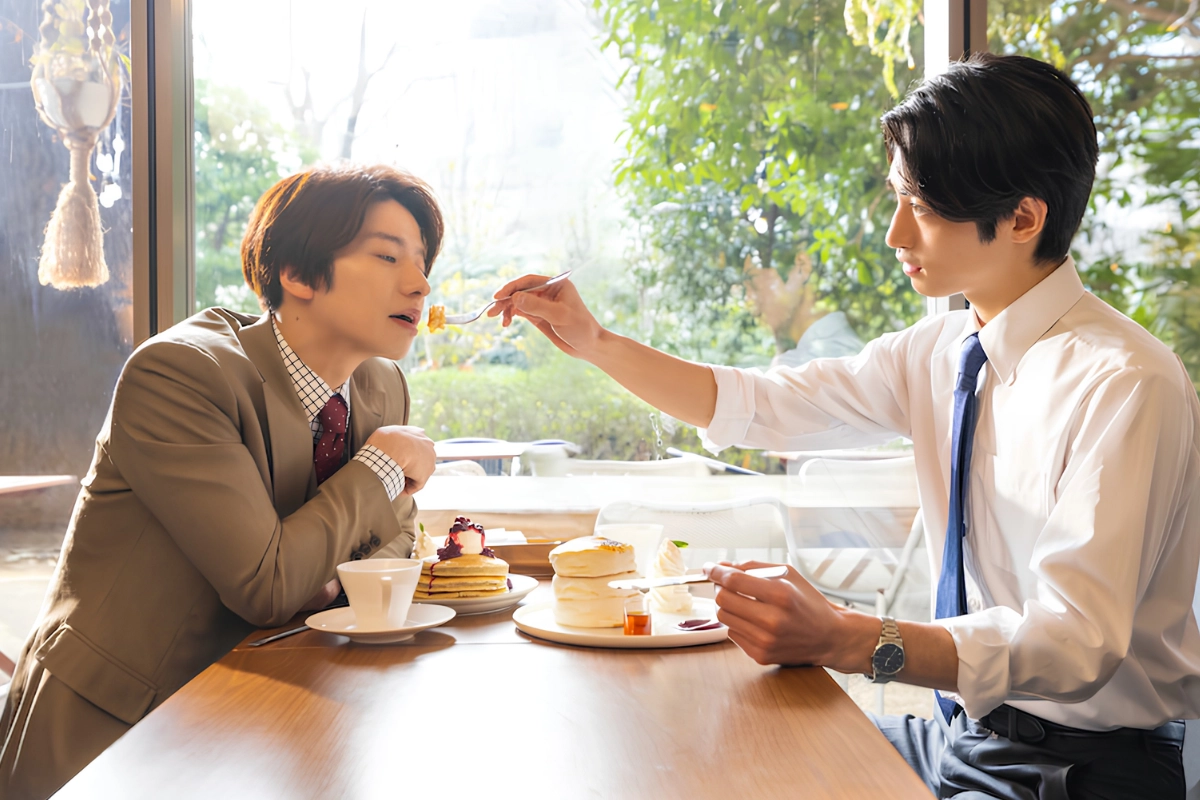 Two Japanese men sit across from each other at a café table, with one feeding the other a bite of dessert.