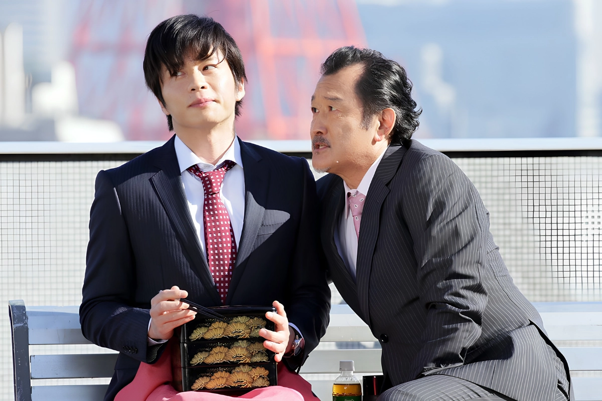 Two Japanese businessmen in suits sit closely together on an outdoor bench, with one leaning toward the other as he holds a bento box.