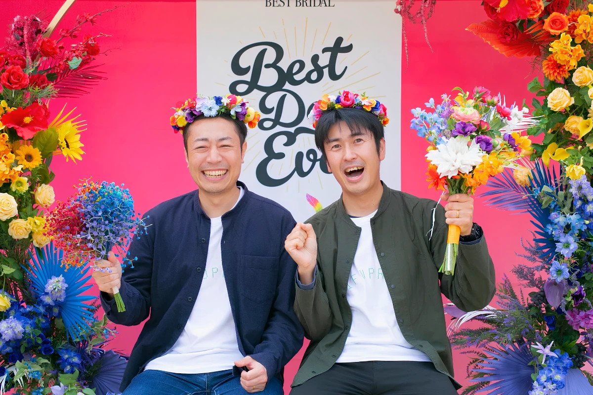 Two smiling men wearing colorful flower crowns pose with bouquets in front of a bright floral Best Bridal photo backdrop.