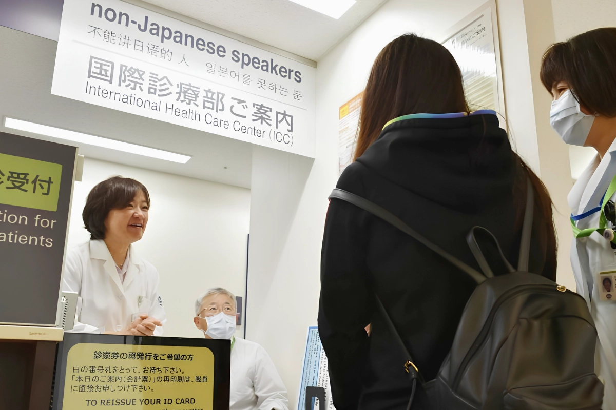 A visitor speaks with hospital staff at an International Health Care Center reception beneath multilingual signage for non-Japanese speakers.